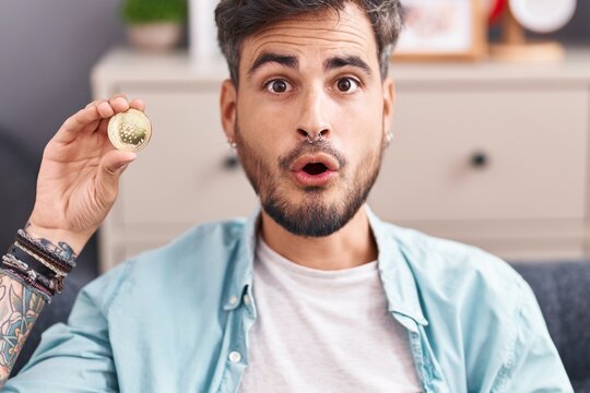Young hispanic man with tattoos holding cardano cryptocurrency coin scared and amazed with open mouth for surprise, disbelief face