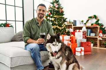 Young hispanic man drinking wine sitting on sofa with dog by christmas tree at home