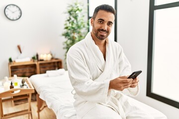 Young hispanic man wearing bathrobe using smartphone at beauty center