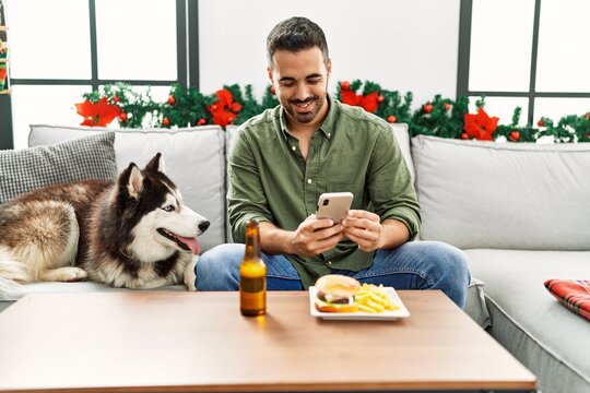 Young hispanic man make photo to hamburger sitting on sofa with dog by christmas decor at home