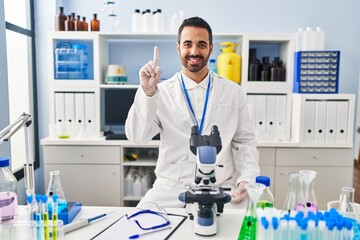 Young hispanic man with beard working at scientist laboratory showing and pointing up with finger number one while smiling confident and happy.