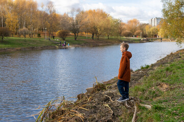 A cute boy stands on the shore of a lake in the park in autumn
