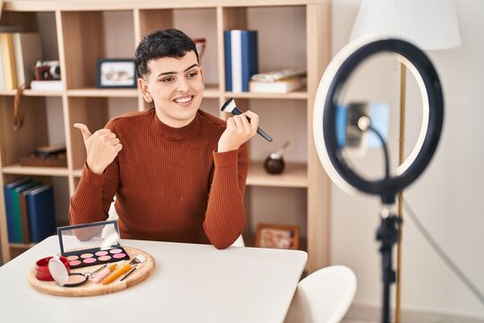Non binary person recording vlog tutorial with smartphone at home pointing thumb up to the side smiling happy with open mouth