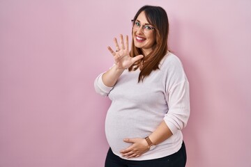 Pregnant woman standing over pink background showing and pointing up with fingers number five while...