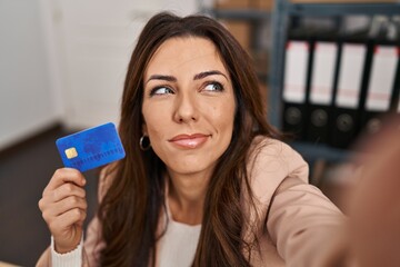 Young brunette woman working at small business ecommerce holding credit card smiling looking to the side and staring away thinking.