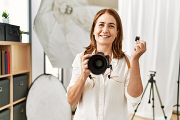 Middle age caucasian woman photographer holding professional camera and memory card at photograph studio
