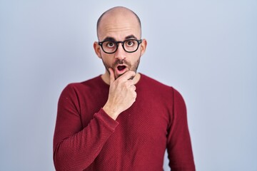 Fototapeta premium Young bald man with beard standing over white background wearing glasses looking fascinated with disbelief, surprise and amazed expression with hands on chin