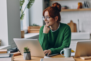 Dreamful young woman using laptop and smiling while working from home