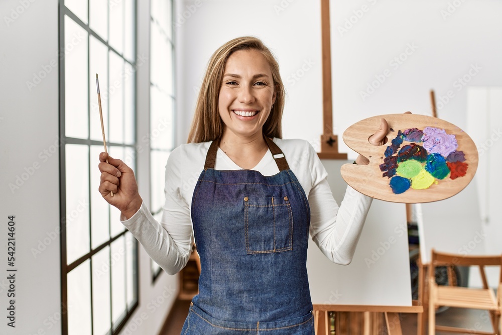 Poster Young caucasian woman smiling confident holding paintbrush and palette at art studio