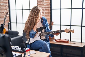Young blonde woman musician playing electrical guitar talking on smartphone at music studio