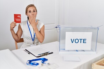 Young blonde woman at political campaign election holding swiss passport covering mouth with hand, shocked and afraid for mistake. surprised expression