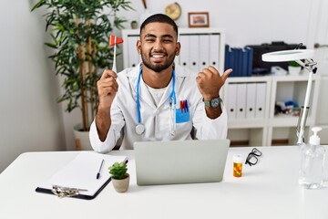 Young indian doctor holding reflex hammer pointing thumb up to the side smiling happy with open...