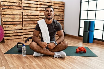 Young indian man sitting on training mat at the gym angry and mad screaming frustrated and furious, shouting with anger. rage and aggressive concept.