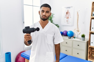 Young indian physiotherapist holding therapy massage gun at wellness center relaxed with serious expression on face. simple and natural looking at the camera.