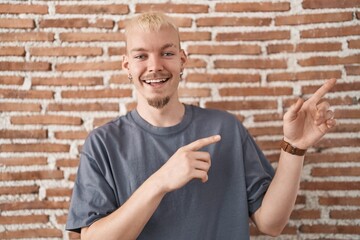 Young caucasian man standing over bricks wall smiling and looking at the camera pointing with two hands and fingers to the side.