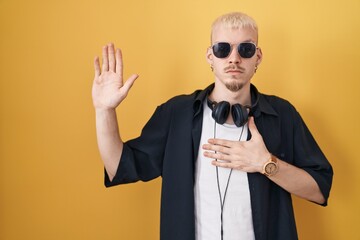Young caucasian man wearing sunglasses standing over yellow background swearing with hand on chest and open palm, making a loyalty promise oath
