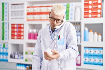 Young caucasian man pharmacist holding pills bottle talking on telephone at pharmacy