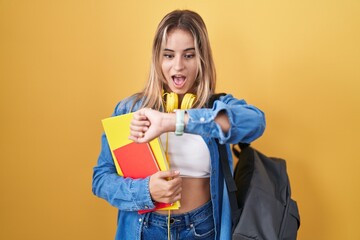 Young blonde woman wearing student backpack and holding books looking at the watch time worried, afraid of getting late