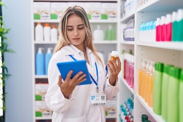 Young blonde woman pharmacist using touchpad holding pills bottle at pharmacy