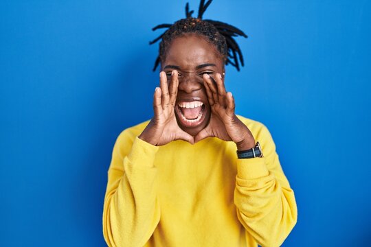 Beautiful black woman standing over blue background shouting angry out loud with hands over mouth