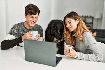 Young caucasian couple using laptop and drinking coffee sitting on the table with dog at home.