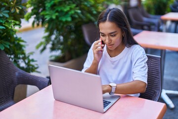 Young beautiful hispanic woman talking on smartphone using laptop sitting on table at coffee shop terrace