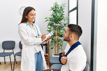 Youg latin injuried man wearing sling talking with the doctor at clinic waiting room.