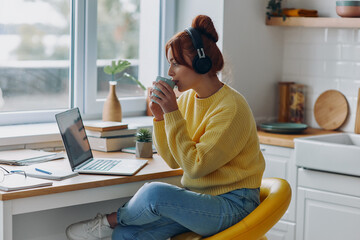 Beautiful woman in headphones drinking coffee and looking at laptop while sitting at the kitchen counter