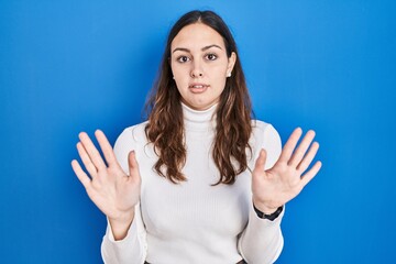 Young hispanic woman standing over blue background moving away hands palms showing refusal and...