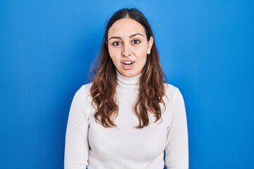 Young hispanic woman standing over blue background afraid and shocked with surprise and amazed expression, fear and excited face.