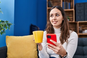 Young beautiful hispanic woman using smartphone drinking coffee at home