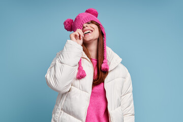 Playful woman in winter coat pulling her funky hat on yes while standing against blue background