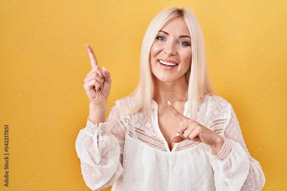 Wall mural Caucasian woman standing over yellow background smiling and looking at the camera pointing with two hands and fingers to the side.