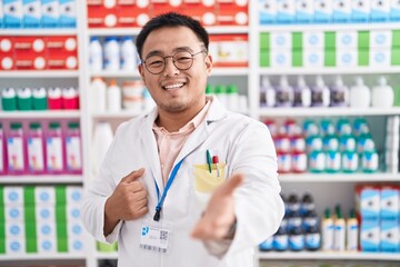 Fototapeta premium Chinese young man working at pharmacy drugstore smiling cheerful offering palm hand giving assistance and acceptance.