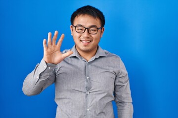 Young chinese man standing over blue background showing and pointing up with fingers number five while smiling confident and happy.