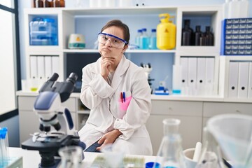 Fototapeta premium Hispanic girl with down syndrome working at scientist laboratory thinking concentrated about doubt with finger on chin and looking up wondering