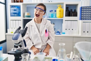 Hispanic girl with down syndrome working at scientist laboratory in shock face, looking skeptical and sarcastic, surprised with open mouth