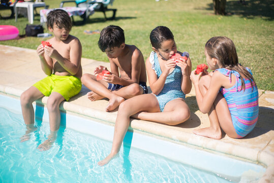 Portrait of four kids eating watermelon on edge of pool. Two little boys and girls in swimwear sitting holding pieces of watermelon talking to each other. Active summer rest, happy childhood concept