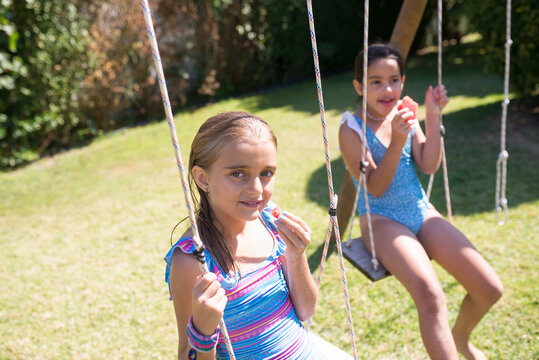 Portrait of two little girls on swings in yard. Happy Caucasian girls in swimsuits resting after swimming sitting on swings and eating pieces of watermelon. Summer vacation and kids leisure concept
