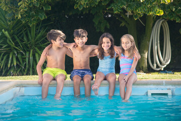 Portrait of happy Caucasian children wetting their legs in pool. Two boys and two girls sitting on edge of pool with legs in blue water laughing and hugging each other. Leisure and friendship concept