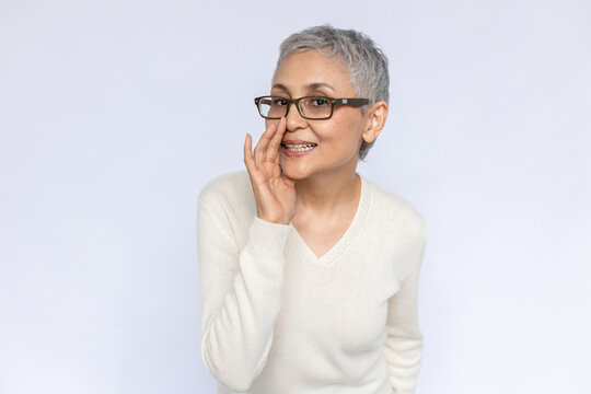 Portrait Of Conspiring Senior Woman Whispering Secret. Mature Caucasian Woman Wearing Eyeglasses And White Jumper Telling Gossip Covering Mouth With Hand. Gossip And Secret Concept