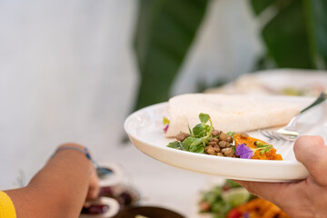 Woman holding a white porcelain plate at the start of a snack. On the plate are green lentil salad, arugula, taco and silver fork.