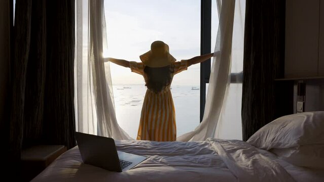 Woman opening white curtains, sitting on bed nearly window,looking to Beautiful sunset above sea, reflection of orange sun in the water after video conference with colleagues, online working on laptop
