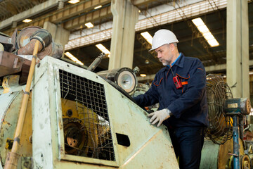 A machine factory engineer is inspecting the machines in the factory with a tablet or laptop and with a deliberate eye check. The factory is big and old.