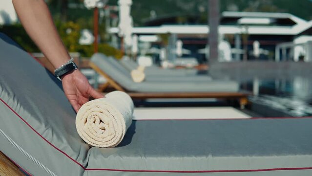 Slow motion close-up of a man laying out a rolled up beige towels on a sun lounger near the pool