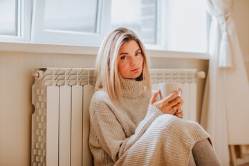 Young blond woman in long winter beige sweater is holding a cup of coffee and posing at home near...