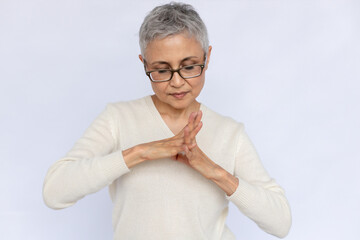 Portrait of tired senior woman stretching fingers over white background. Mature Caucasian woman wearing eyeglasses and white jumper having problems with joints. Aging diseases concept