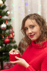 Young beautiful woman in a red sweater with a mug of cocoa and marshmallows at home by the new year tree on new year's eve. Selective focus. Portrait