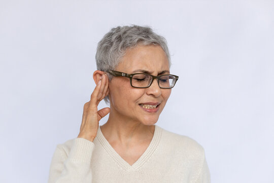 Portrait of irritated senior woman suffering from eardrum. Mature Caucasian woman wearing eyeglasses and white jumper touching ear, annoyed with noise. Earache concept