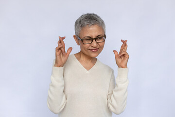 Portrait of deceitful senior woman making liar gesture. Mature Caucasian woman wearing eyeglasses and white jumper crossing fingers in hope over white background. Liar, hope, wish concept